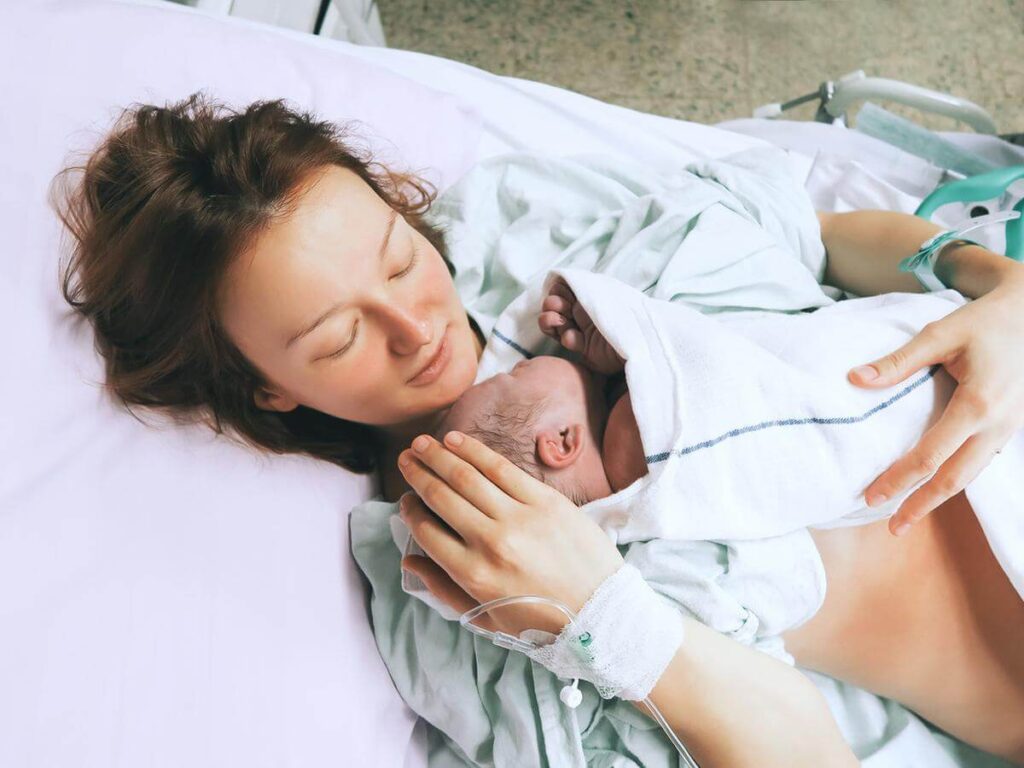 Kobieta leżąca na szpitalnym łóżku, obejmująca nowo narodzone niemowlę owinięte w biały kocyk