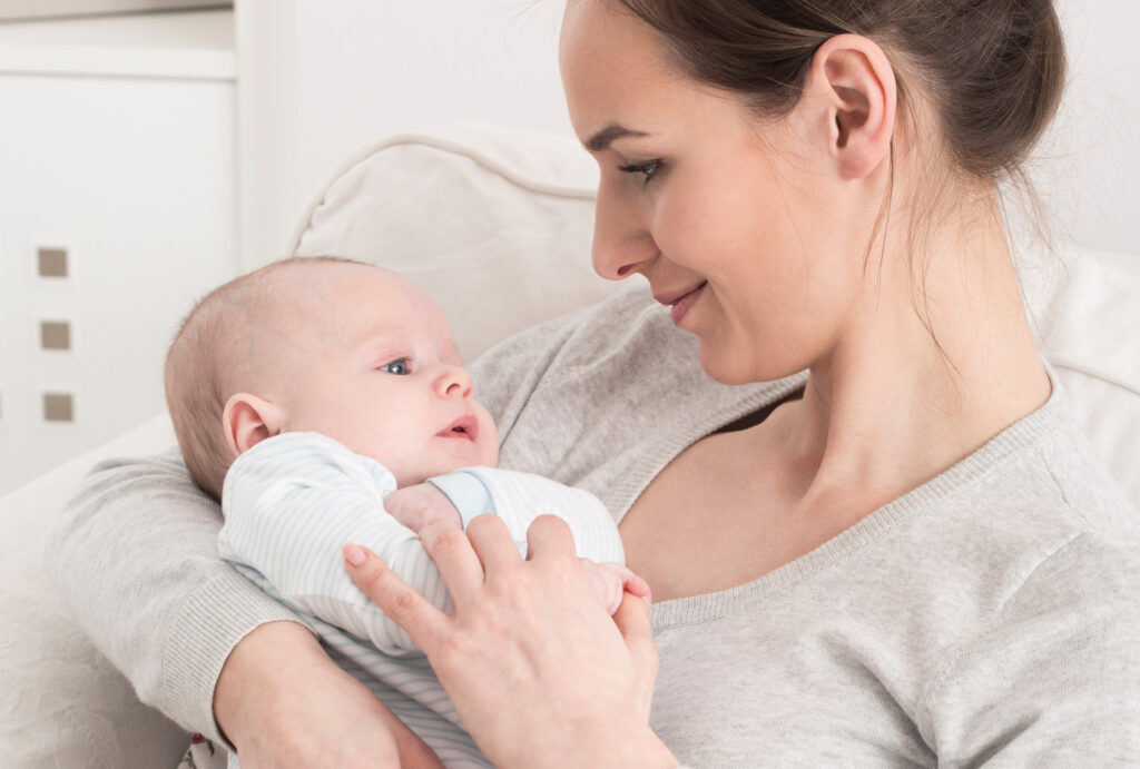 Kobieta trzymająca niemowlę w ramionach, ujęcie w jasnym pomieszczeniu, oboje patrzą na siebie