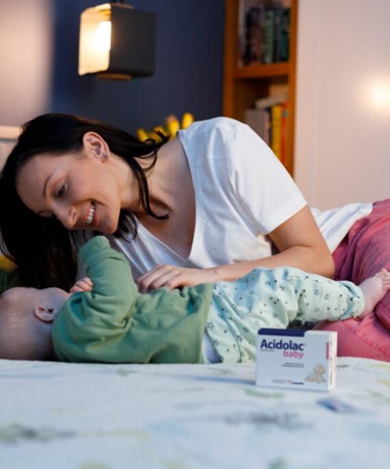 Mama z niemowlęciem na łóżku, obok opakowania Acidolac Baby